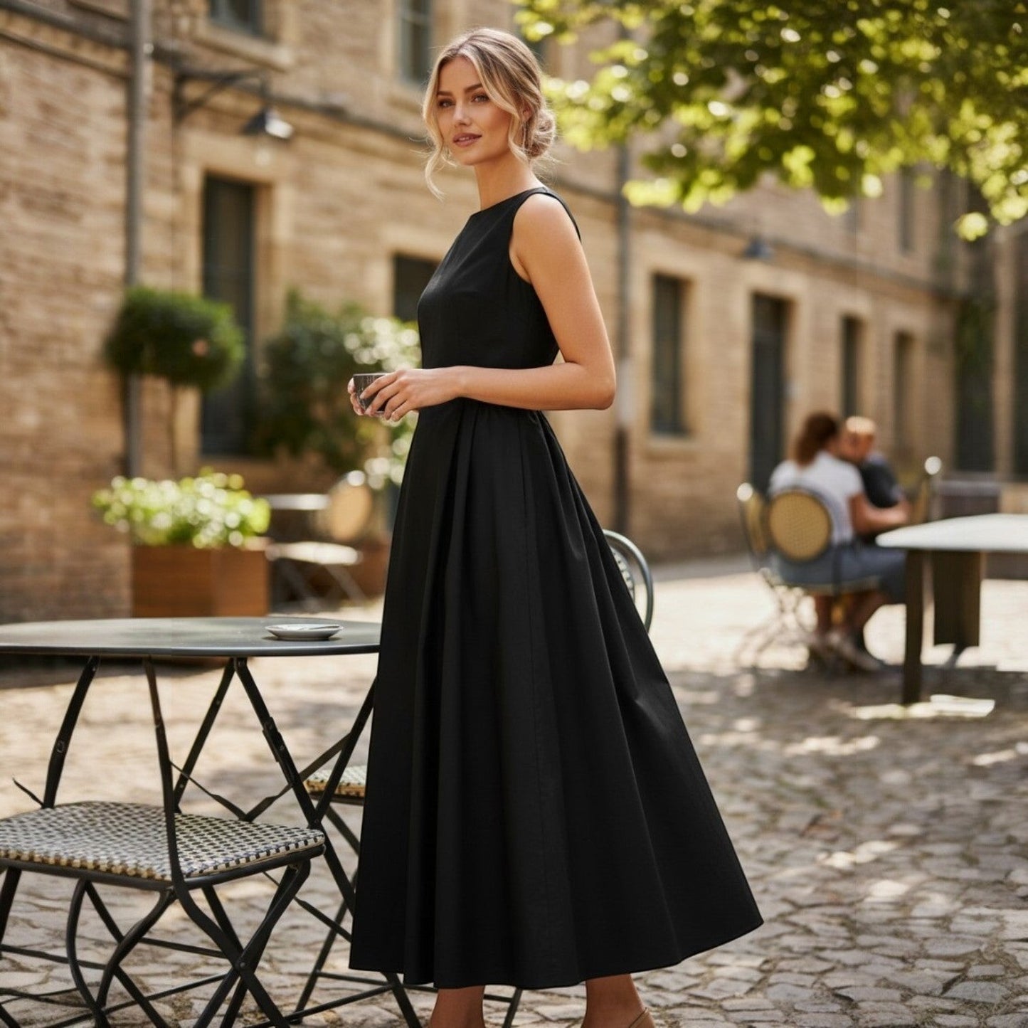La Vie Continue black 100% cotton A-line maxi dress with a boat neckline, sleeveless design, and a full skirt. Model stands outdoors at a cafe.