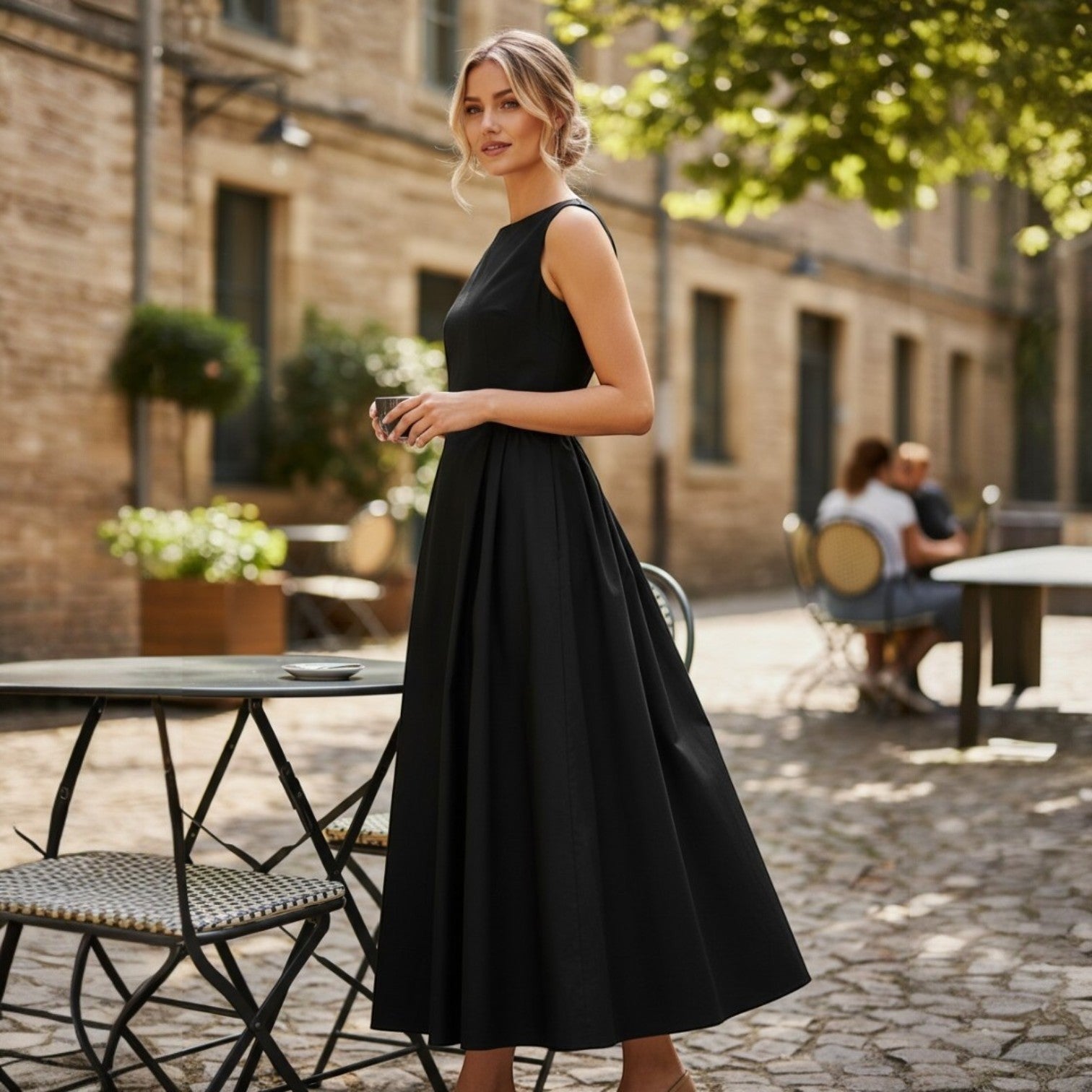 La Vie Continue black 100% cotton A-line maxi dress with a boat neckline, sleeveless design, and a full skirt. Model stands outdoors at a cafe.